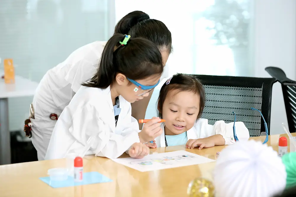 Dva otroka in ženska v laboratorijskem plašču barvajo sliko za mizo