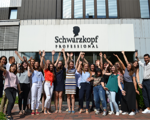 Raznolika Henklova ekipa stoji in navija pred poslovno stavbo Schwarzkopf in dviguje roke