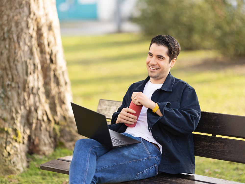 Henklov uslužbenec sedi na klopi v parku in dela na prenosnem računalniku. 