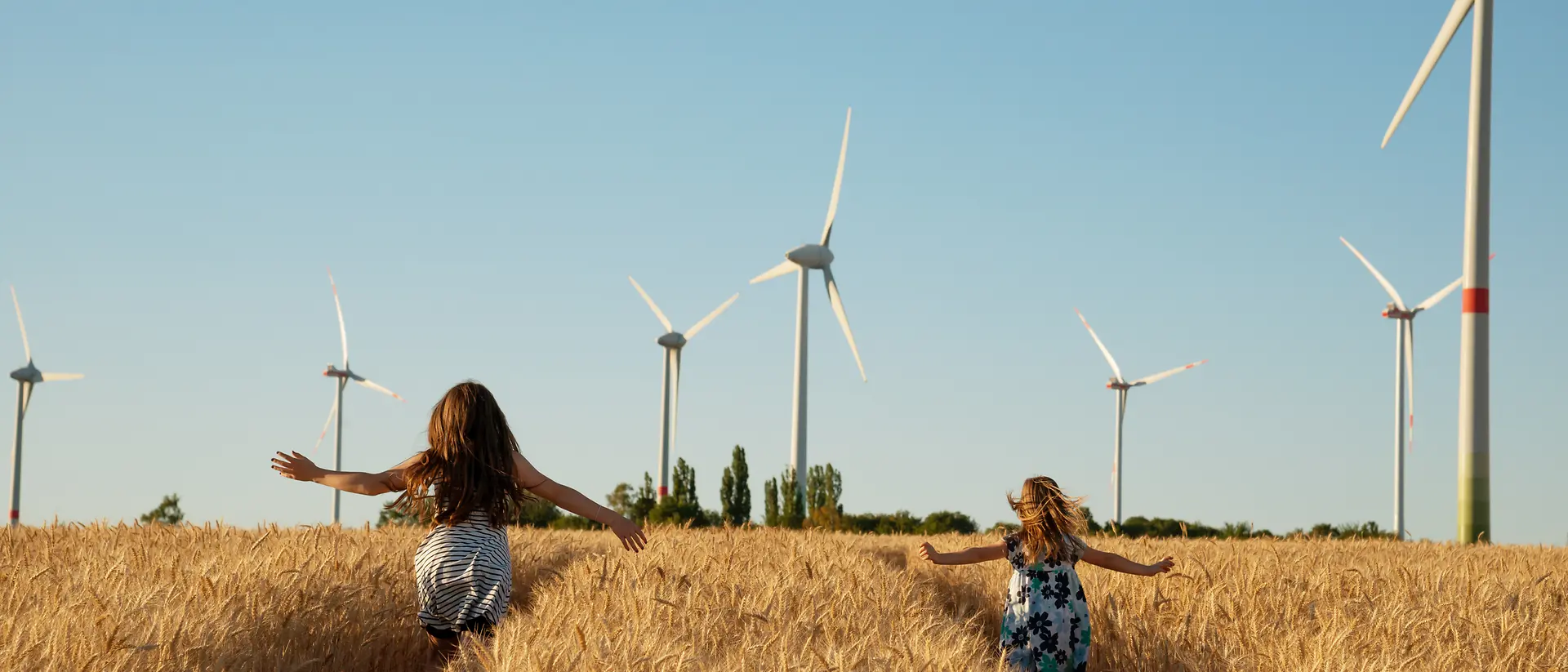 Dve dekleti, ki tečeta po koruznem polju s sodobnimi mlini na veter v ozadju