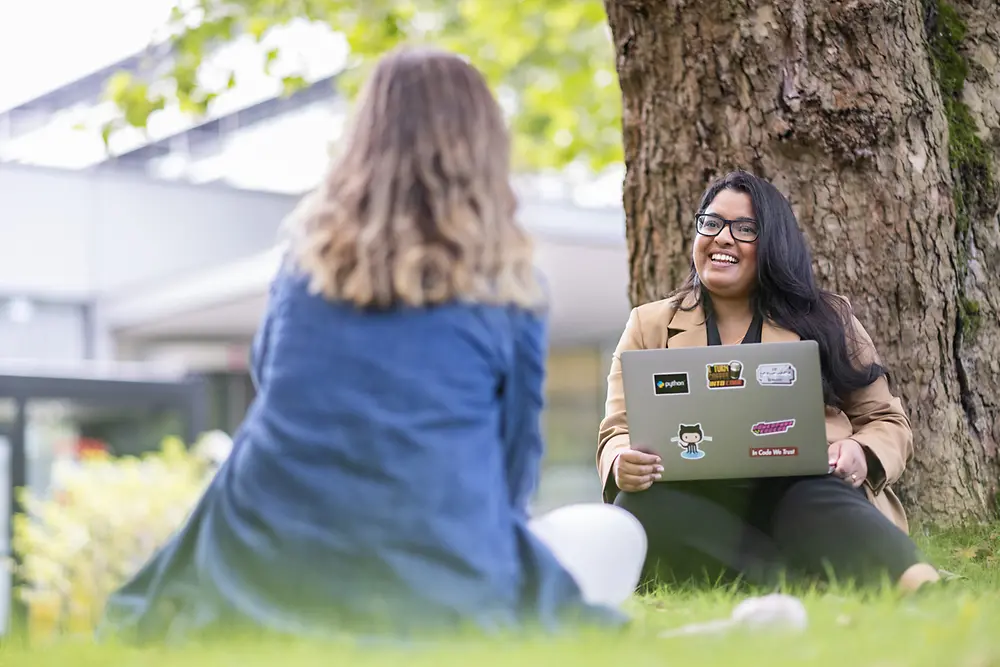 Two women are talking sitting on a meadow, one of them is holding a laptop in her hand.