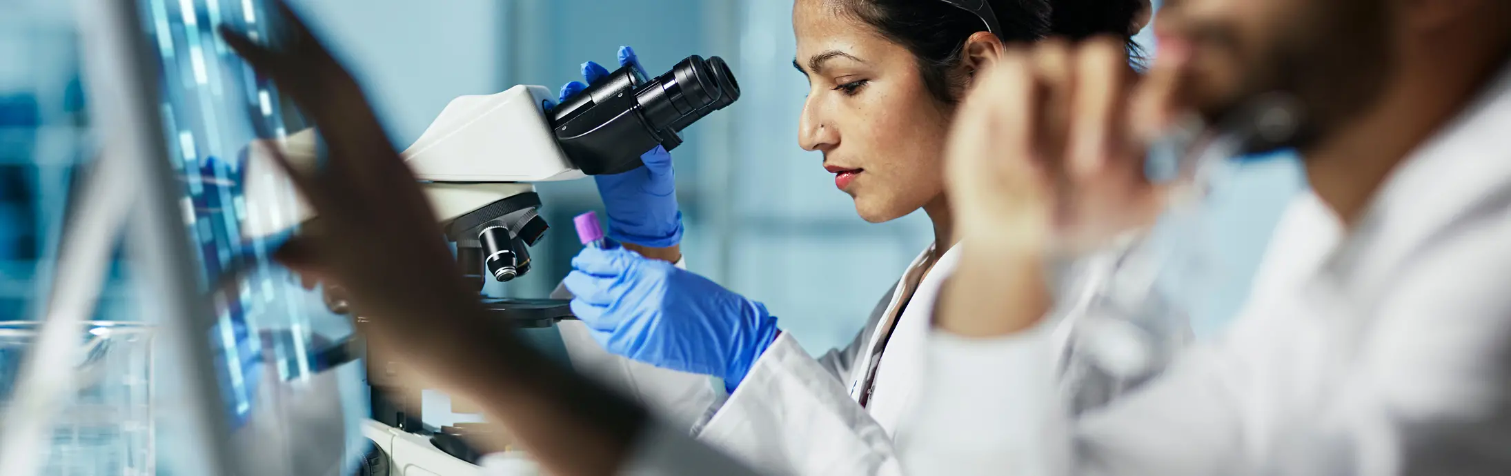 Laboratorij z žensko, ki sedi pred mikroskopom in si ogleduje vzorec, in moškim z brado spredaj, ki gleda v zaslon 