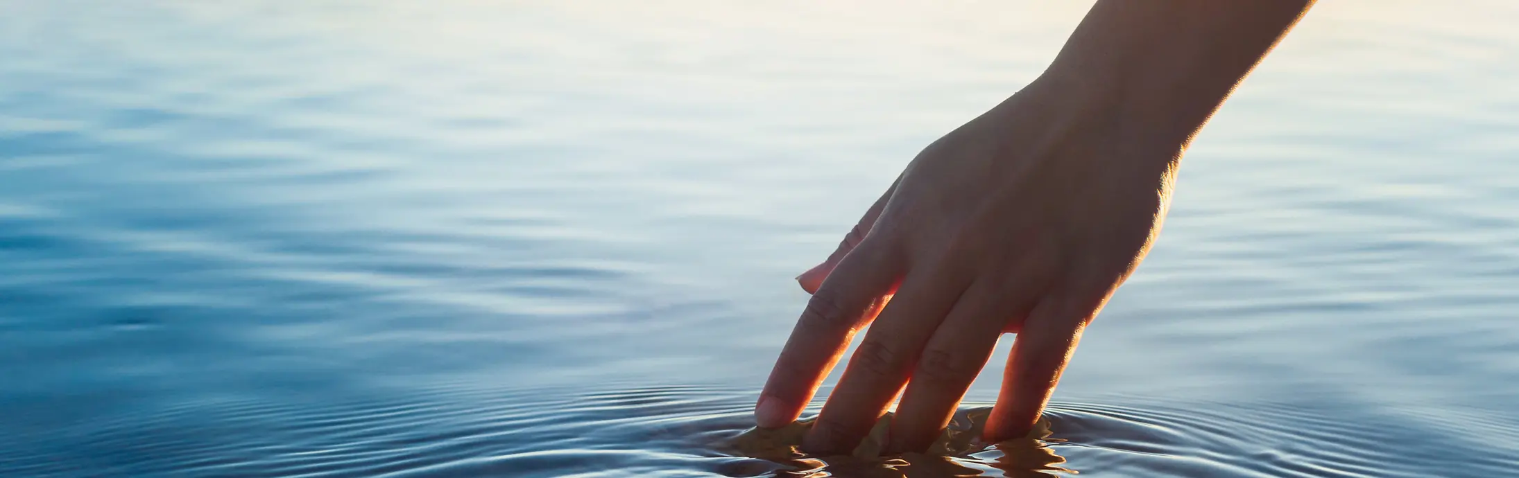 Roka se dotika mirne vodne gladine pred obzorjem