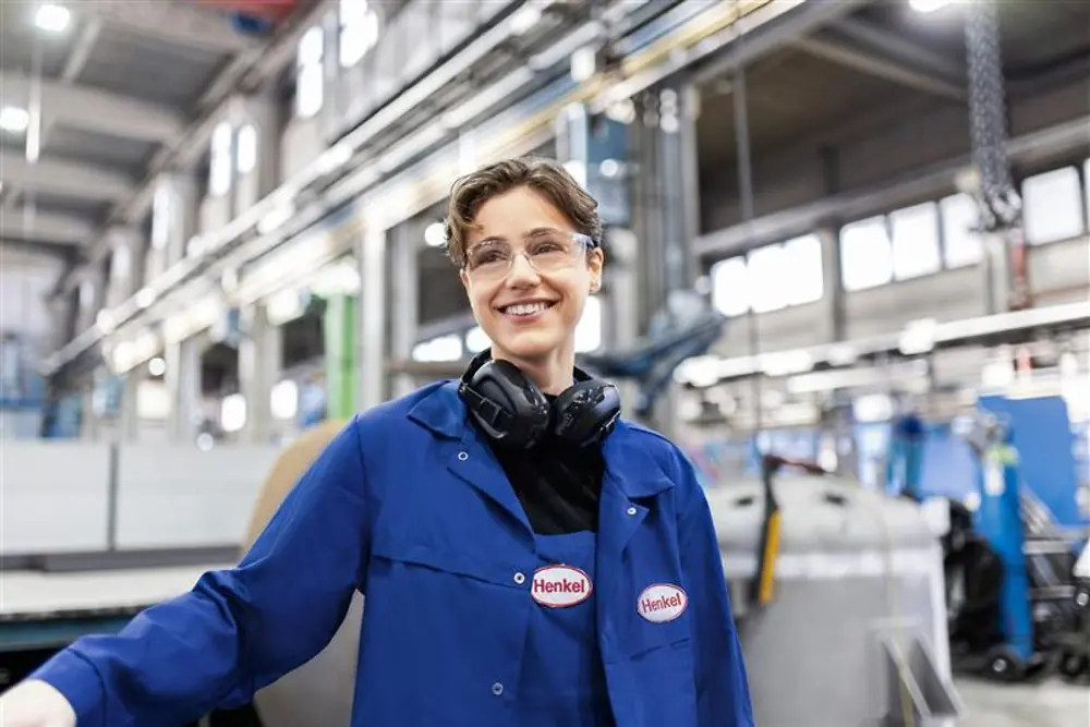 Nasmejani Henklov zaposleni v modri delovni halji in z zaščitnimi slušalkami stoji v industrijski proizvodni hali in predstavlja strokovno znanje in inovacije v proizvodnji.
