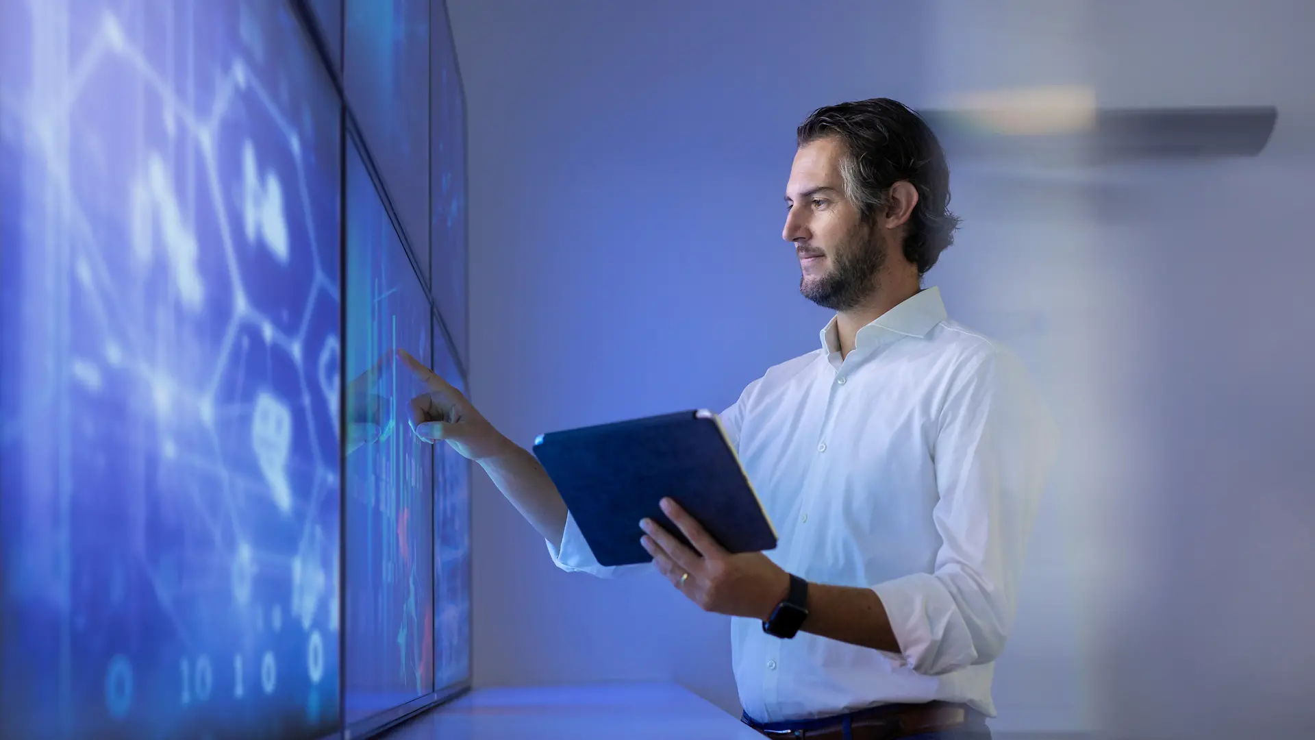 Moški stoji poleg digitalnih zaslonov in s prstom kaže na zaslon, medtem ko drži iPad