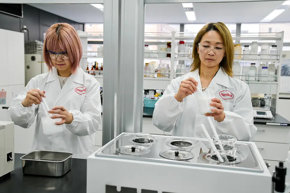 Dva posameznika, ki nosita bele Henklove laboratorijske plašče, delata v sodobnem laboratoriju. Na pultu rokujejo z znanstvenimi instrumenti in posodami, v ozadju pa je vidna različna laboratorijska oprema in police z zalogami.